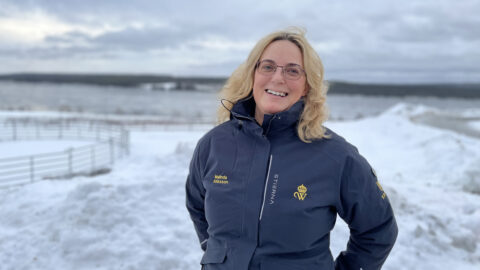 Malinda Alriksson, i blå jacka, står och ler i ett snölandskap med en frusen sjö i bakgrunden.