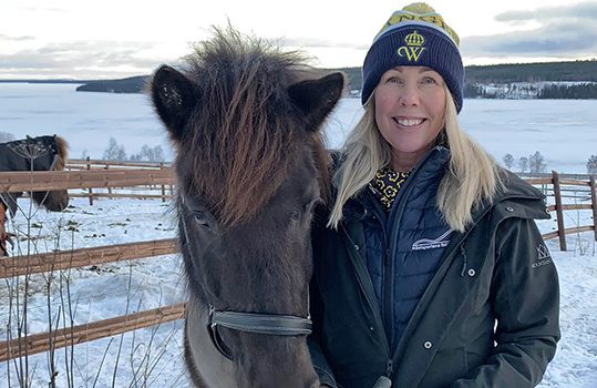 Blond kvinna i gul och blå mössa med ett Wångenemblem på tittar in i kameran och ler. Bredvid henne står en svart islandshäst i grimma. I bakgrunden syns en häst med täcke i en hage.