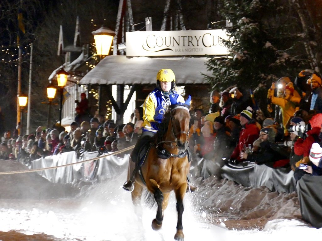 Varmblodshäst galopperar så snön ryker, ryttare bär Wångens gula och blå kläder. I bakgrunden syns publik till skijoring-tävling och skylt med texten Country Club.