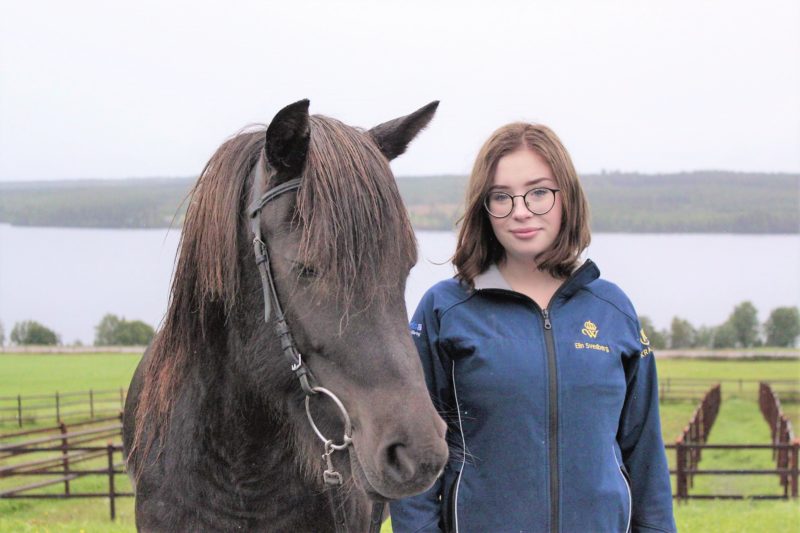 Svart islandshäst i träns står bredvid Elin Svedberg. Elin har mörkt pageklippt hår, runda glasögon och jacka med Wångens emblem. I bakgrunden syns hagar och Alsensjön