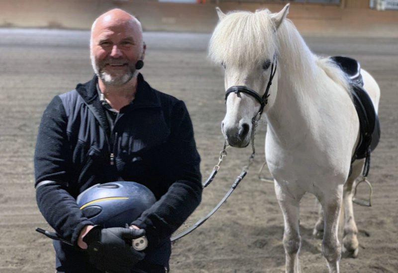 Man med mikrofonheadset ler mot kameran. Han håller hjälmen i handen och tyglarna till en vit häst som står bakom honom