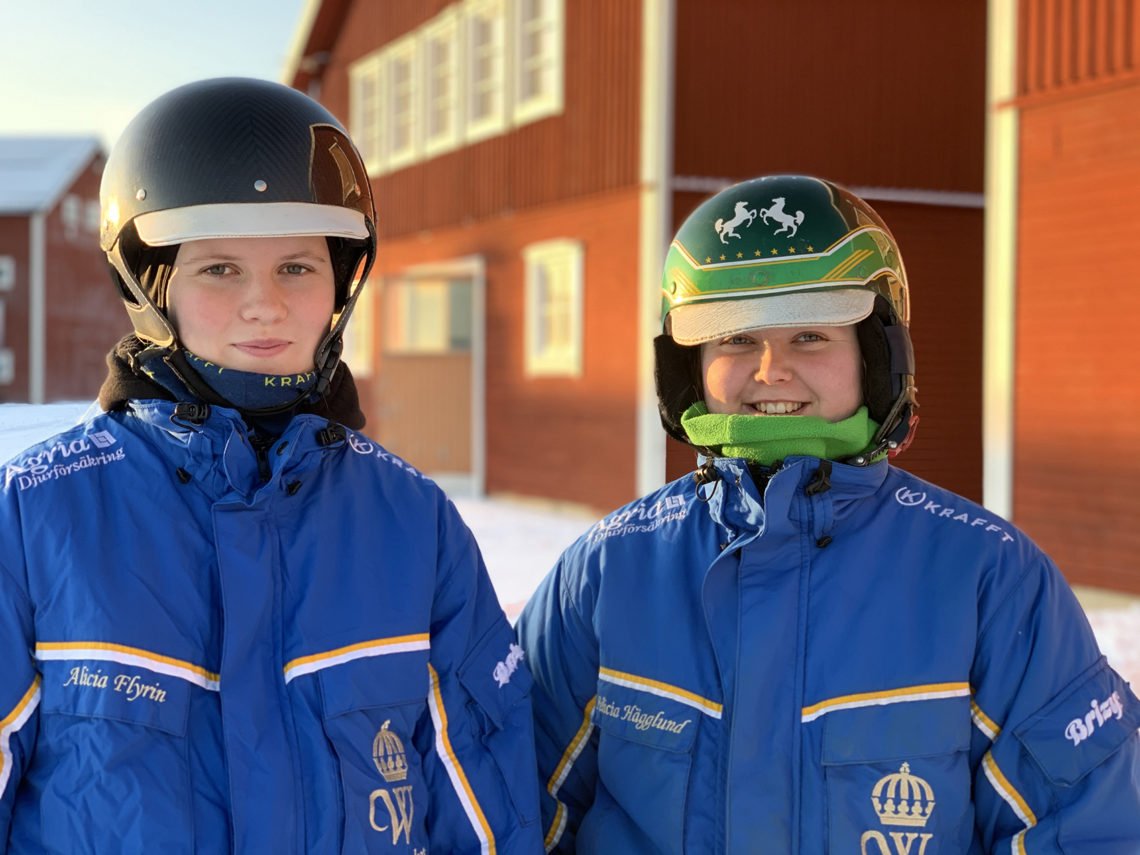 Två studenter på hippologprogrammet trav står i travdressar och travhjälmar står framför röda stallar