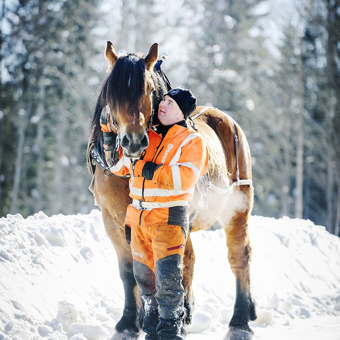 Man i varselkläder och mössa står bredvid och håller i sin häst i ett snöigt skogslandskap. Det är kallt, utandningsluften syns från hästen