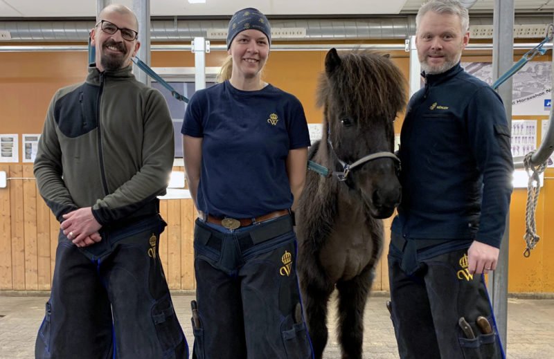 Emanuel, Johanna och Hjalti står i skohallen iförda hovslagarförkläden med Wångens sigill på. Mellan dem står en svart islandshäst. Det är de tre lärarna på hovslagarutbildningen.