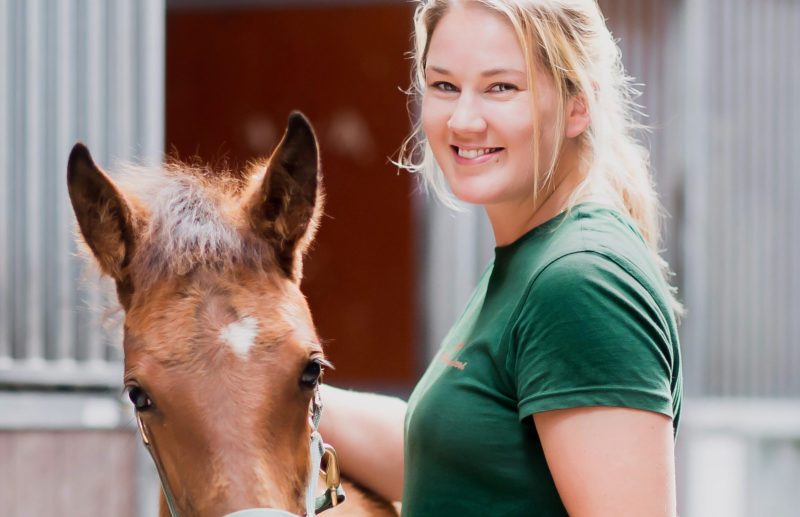 Kvinna tittar in i kameran ler hon håller ett föl bredvid sig i grimma