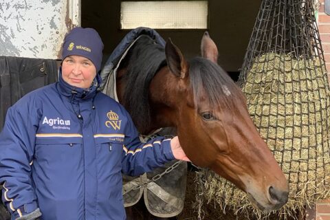 Tina står utanför en box på träningsanläggningen Grosbois i Frankrike. Hon har en blå Wången-mössa och en blå Wångenjacka på sig. En varmblodstravare tittar ut ur boxen.