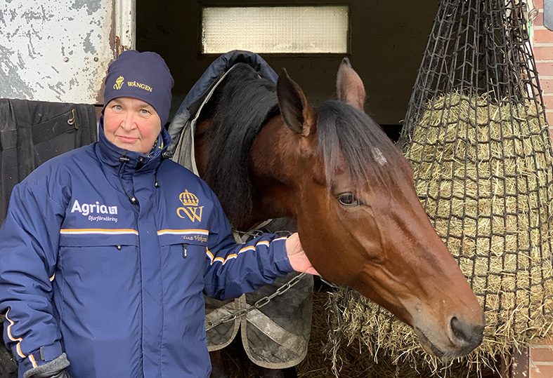 Tina står utanför en box på träningsanläggningen Grosbois i Frankrike. Hon har en blå Wången-mössa och en blå Wångenjacka på sig. En varmblodstravare tittar ut ur boxen.