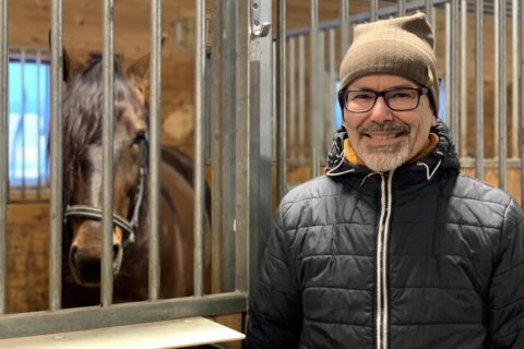 En man i förd jacka mössa och svartbågade glasögon ler in i kameran. Han står utanför en box i ett stall. I boxen står en varmblodstravare