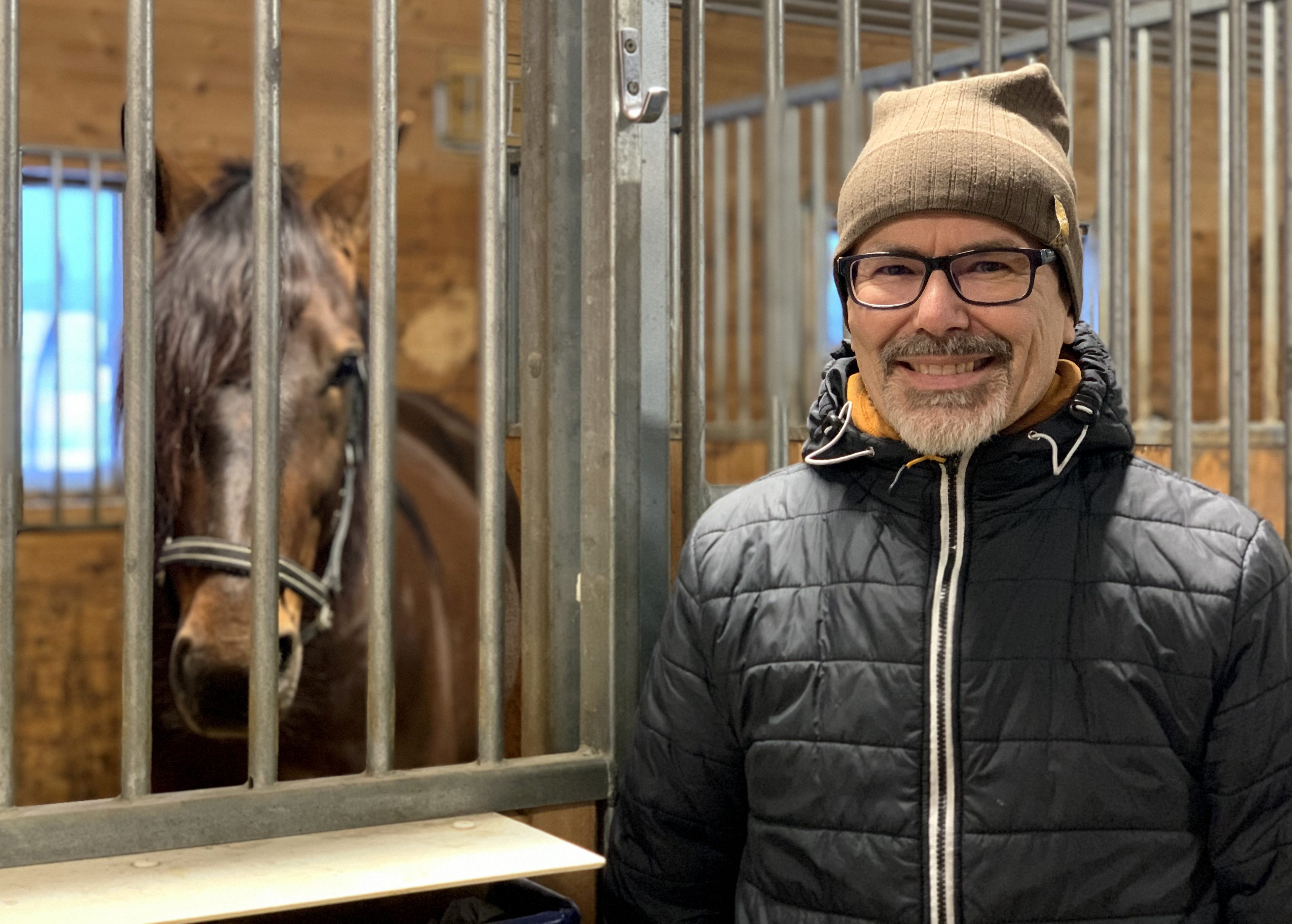 En man i förd jacka mössa och svartbågade glasögon ler in i kameran. Han står utanför en box i ett stall. I boxen står en varmblodstravare