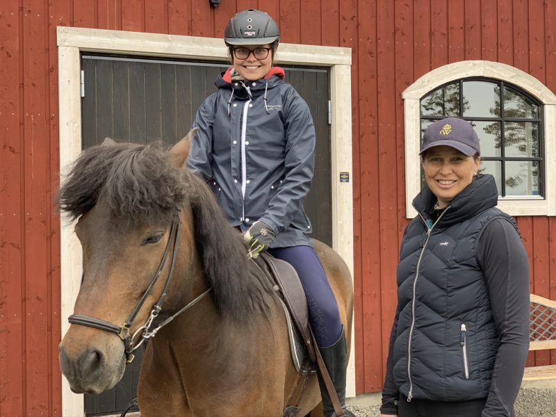 Flicka rider på brun islandshäst och kvinna står bredvid på marken. Bakom dem syns ett rött stall