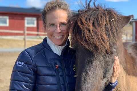 Malin Appelqvist står i en hage tillsammans med en islandshäst. Hon har en blå täckjacka med Wångens sigill på och ler in i kameran. Det är en soligt vårig dag.