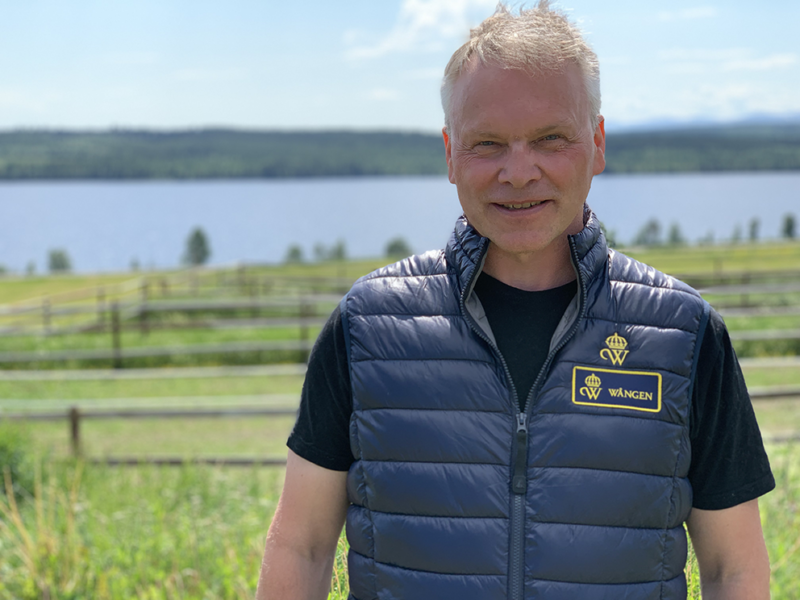 Guðni Ágústsson står utomhus på Wången en sommardag. Sjön syns i bakgrunden och han har en väst som det står Wången på