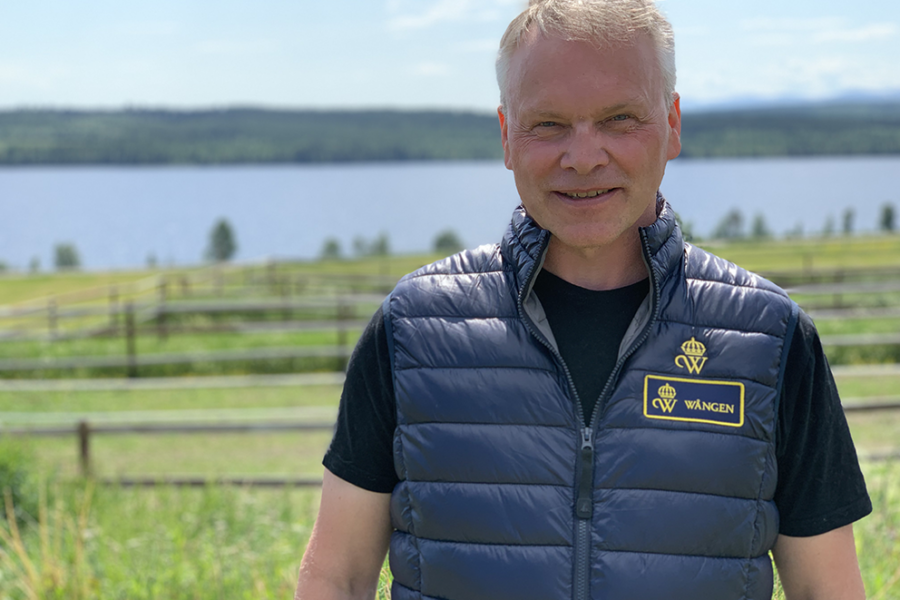 Guðni Ágústsson står utomhus på Wången en sommardag. Sjön syns i bakgrunden och han har en väst som det står Wången på