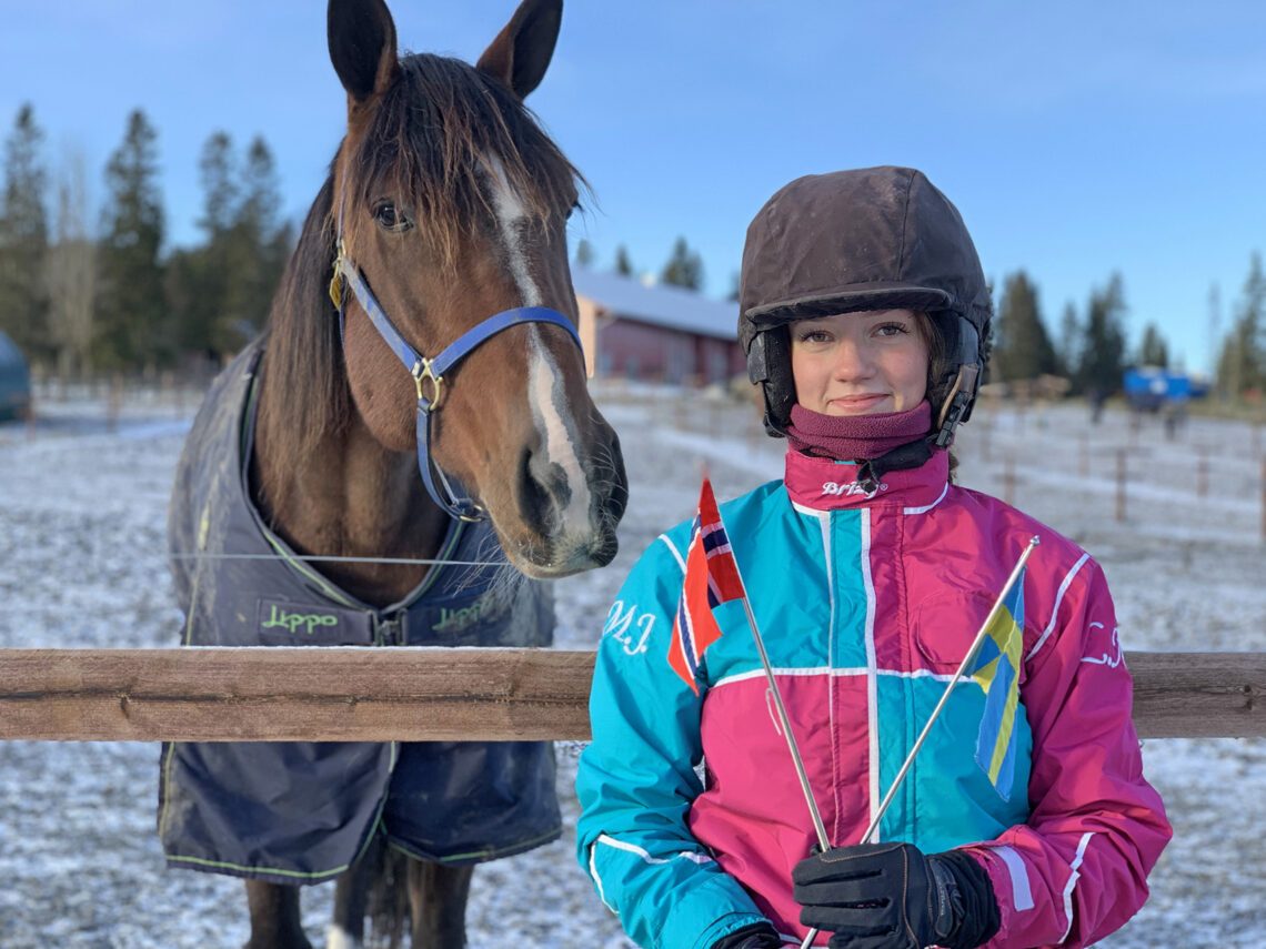 En varmblodstravare står i en hage och tittar ut över stängslet. Framför står en tjej i svart hjälm och färgglad kuskdress. I händerna håller hon en svensk och en norsk flagga inför ETSÄ-loppet.