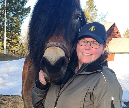 Jennie Runesson i en blå keps med Wångens sigill på ler in i kameran. En nordsvensk brukshäst i grimma står bredvid henne.