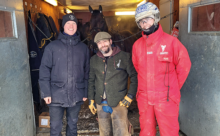 Tre män står i en stalldörr i vinterkläder. Inne i stallet syns en varmblodstravare uppställd på stallgången.