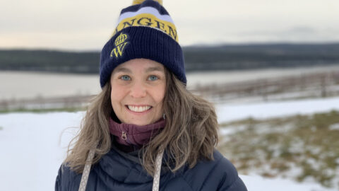 Karin Augustyniak i marin vinterjacka och Wångenmössa, står omgiven av snödammad mark och en naturskön bakgrund av vatten och kullar.