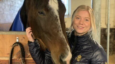 Amanda Lundell står i stallet utanför en box och klappar en häst.