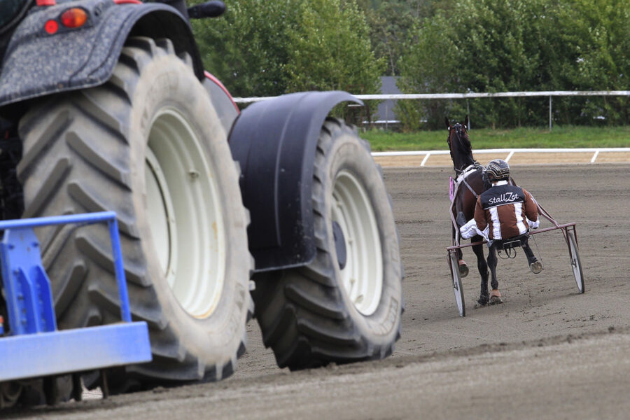 En varmblodstravare i sulky körs av en kusk med texten Stall Zet på. Bakom ekipaget syns en stor traktor framträdande i förgrunden. Bilden är från Jägersros travbana.