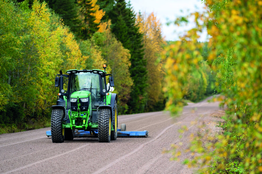 En grön traktor drar ett blått redskap på en grusväg omgiven av gröna och gula träd, som skulle kunna vara en scen från anläggningsprofilen.