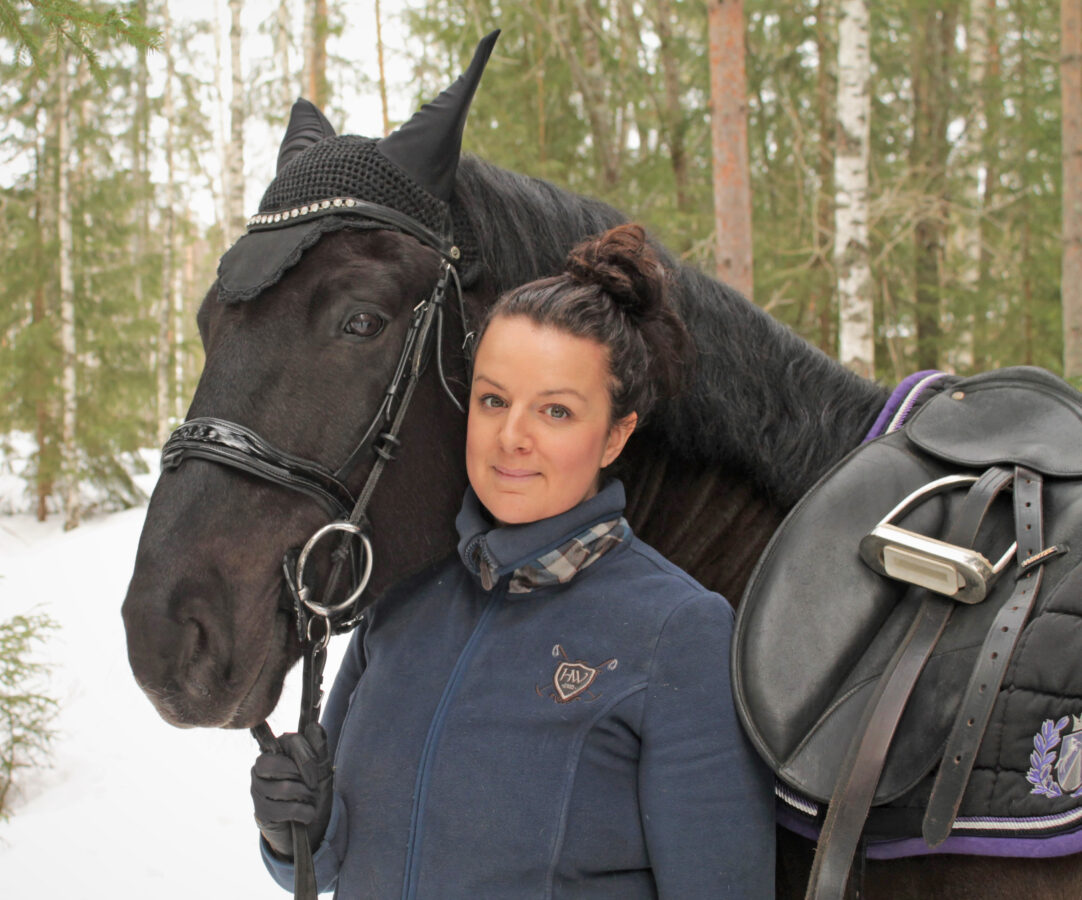 Julia Bergquist står bredvid en svart ridhäst i en snöig skog.