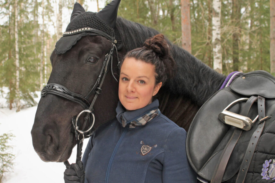 Julia Bergquist står bredvid en svart ridhäst i en snöig skog.