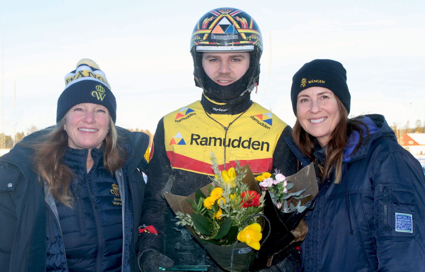 Mats Djuse står i kuskdress och hjälm. I handen har han blommor och en glasstatyett med texten Wångens guldstjärna ingraverad. Bredvid honom på var sida står två kvinnor i blå jackor och Wångenmössor.