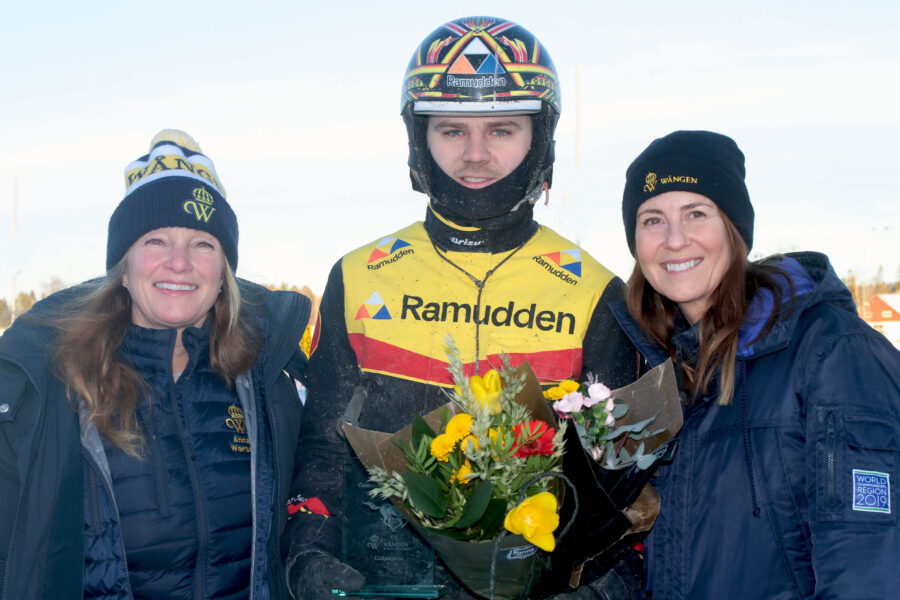 Mats Djuse står i kuskdress och hjälm. I handen har han blommor och en glasstatyett med texten Wångens guldstjärna ingraverad. Bredvid honom på var sida står två kvinnor i blå jackor och Wångenmössor.