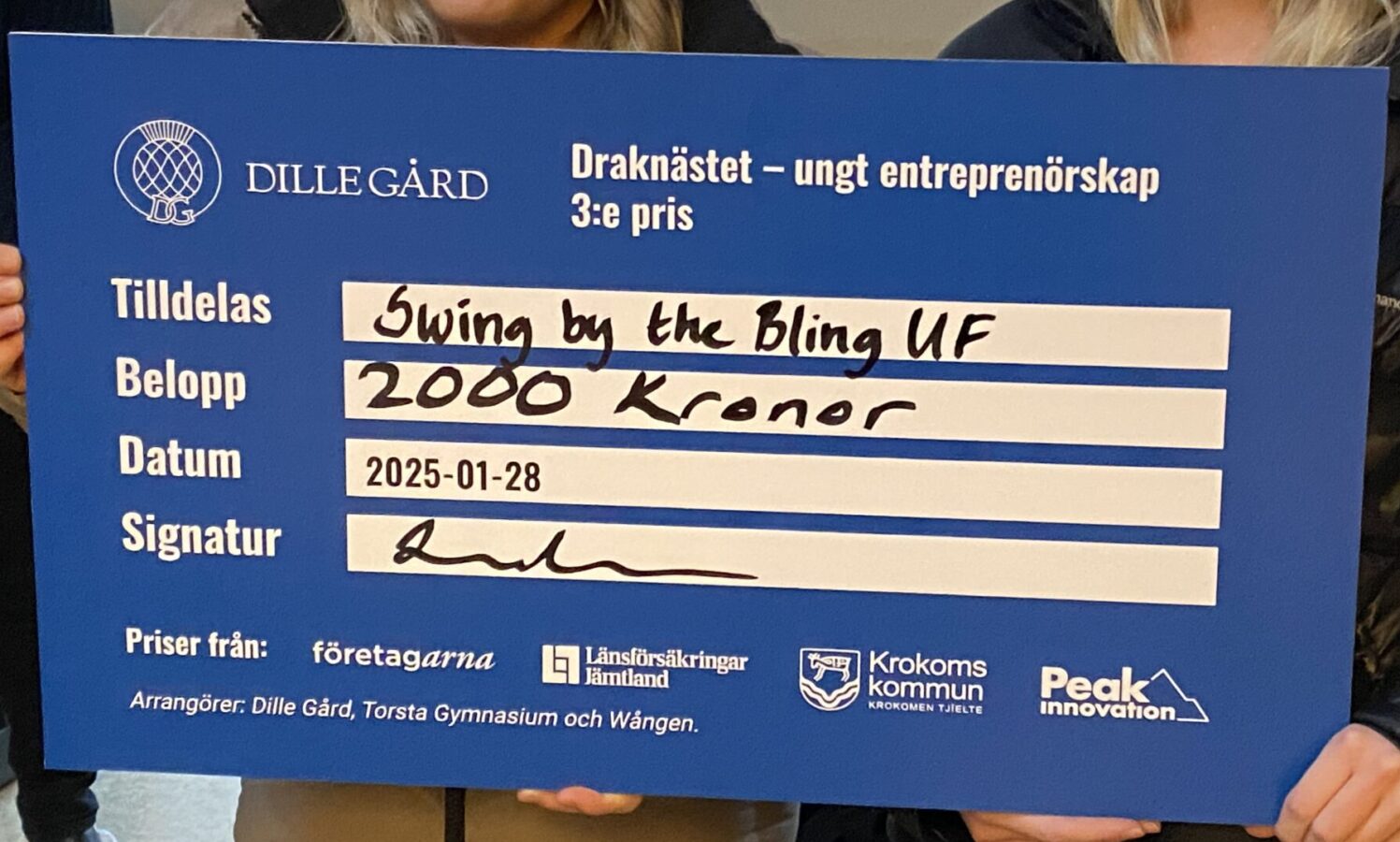 Två UF-företagare håller stolta upp en stor blå check för "Swing by the Bling UF", tilldelad 2000 kr sponsrat av Krokoms kommun, Företagarna, Länsförsäkringar Jämtland och Peak Innovation