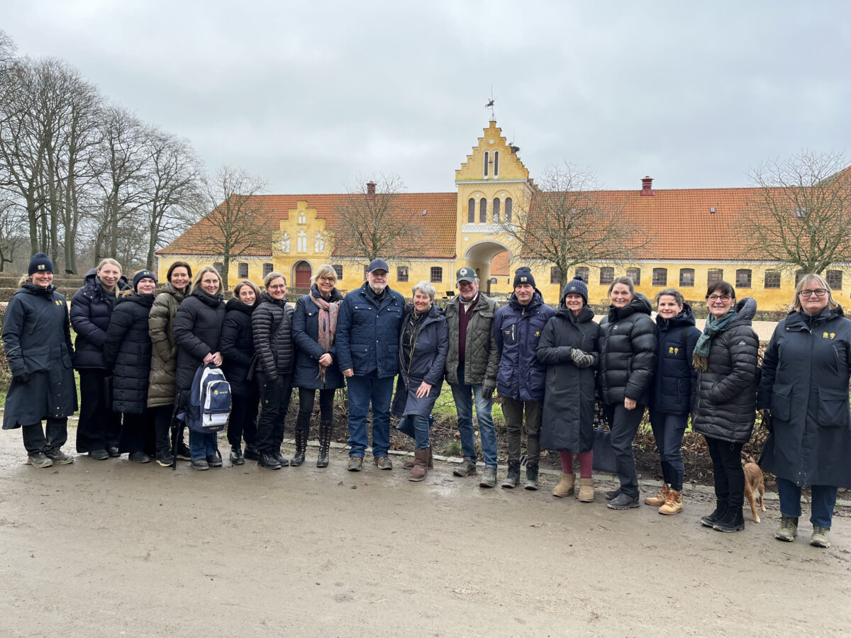 En grupp klädda i vinterkläder, står uppradade utomhus framför en stor gul byggnad med rött tak en molnig dag.