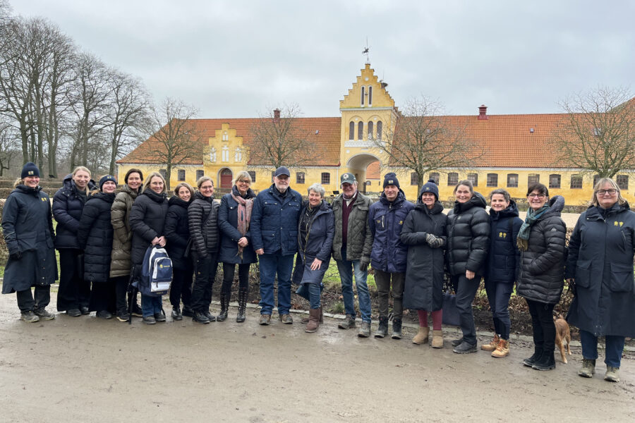 En grupp klädda i vinterkläder, står uppradade utomhus framför en stor gul byggnad med rött tak en molnig dag.