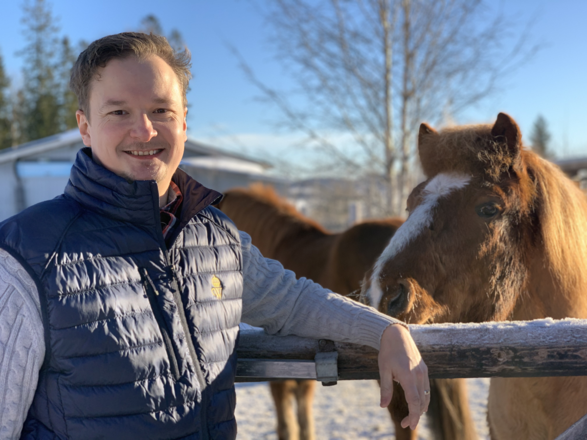 Jens Hernerud står i en blå väst vid ett trästaket. Två islandshästar syns i bakgrunden, den ena nosar nyfiket mot Jens. Det är en vacker vinterdag.