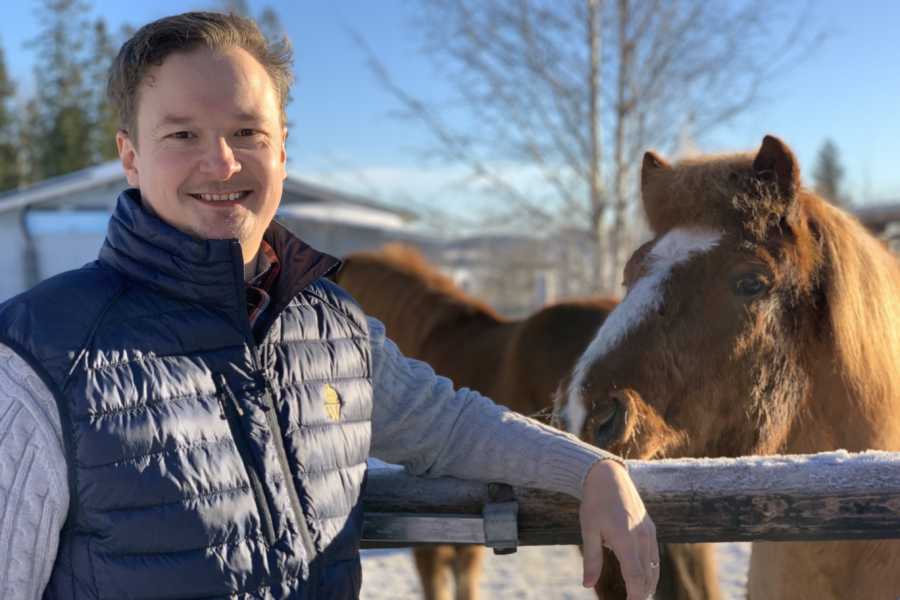 Jens Hernerud står i en blå väst vid ett trästaket. Två islandshästar syns i bakgrunden, den ena nosar nyfiket mot Jens. Det är en vacker vinterdag.