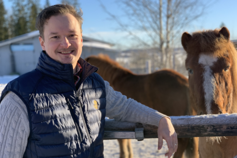 Jens Hernerud står i en blå väst vid ett trästaket. Två islandshästar syns i bakgrunden, den ena tittar nyfiket in i bild. Det är en vacker vinterdag.