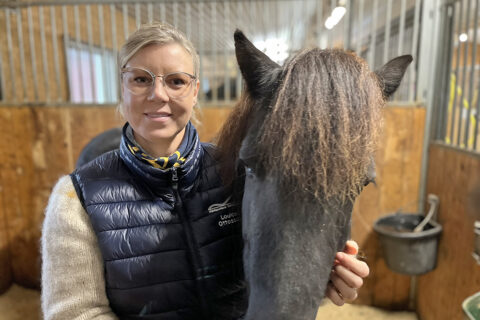 Louice Ottosson står i en box tillsammans med en svart islandshäst. Hon är blond och har glasögon.