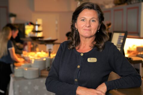 Lisa Eliasson står i restaurangen med armarna vilande på disken. I bakgrunden tar två personer för sig en buffé.