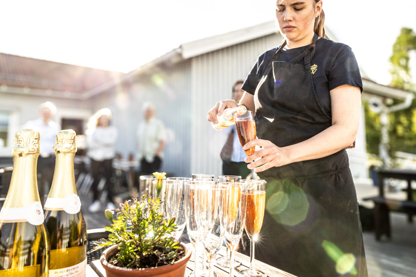 En kvinna i förkläde häller elegant upp champagne i glas på ett utomhusbord prytt med flaskor. I bakgrunden pratar flera personer och njuter av solen.