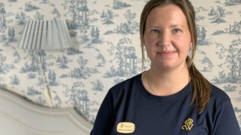 En person i marinblå t-shirt står i ett rum med mönstrad tapet. En namnskylt med texten "Rebecka Erlandsson" syns och en lampa lyser mjukt i bakgrunden.