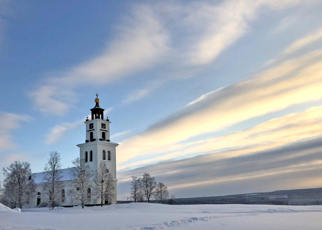 En vit kyrka med ett högt torn står i ett snölandskap under en delvis molnig himmel.