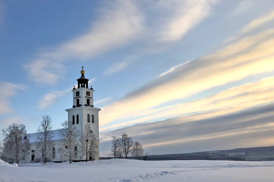 En vit kyrka med ett högt torn står i ett snölandskap under en delvis molnig himmel.