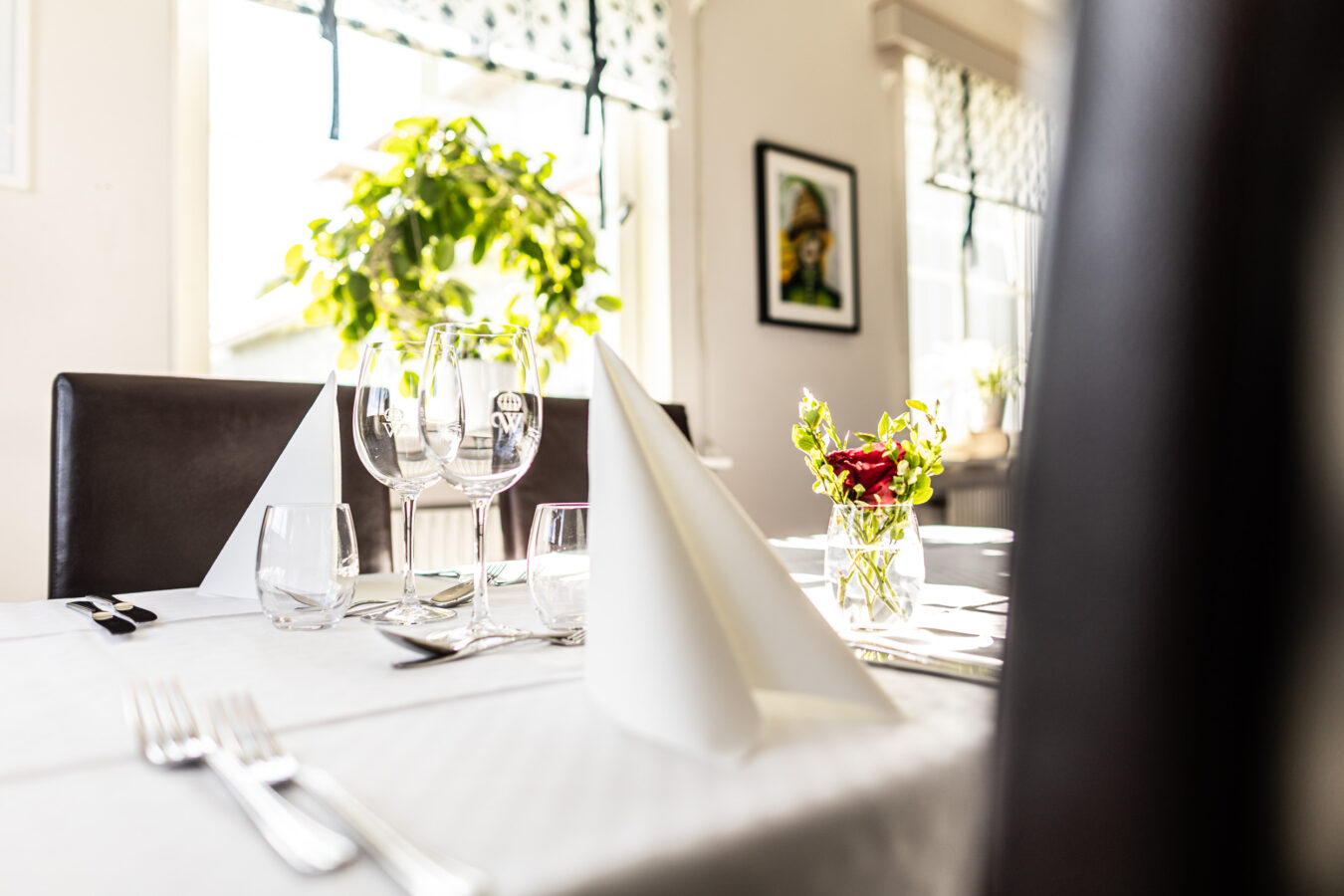 Elegant matbordsuppställning med vikta servetter, glas och en liten blomvas. Ljust rum med en inramad bild på väggen och en planta vid fönstret.