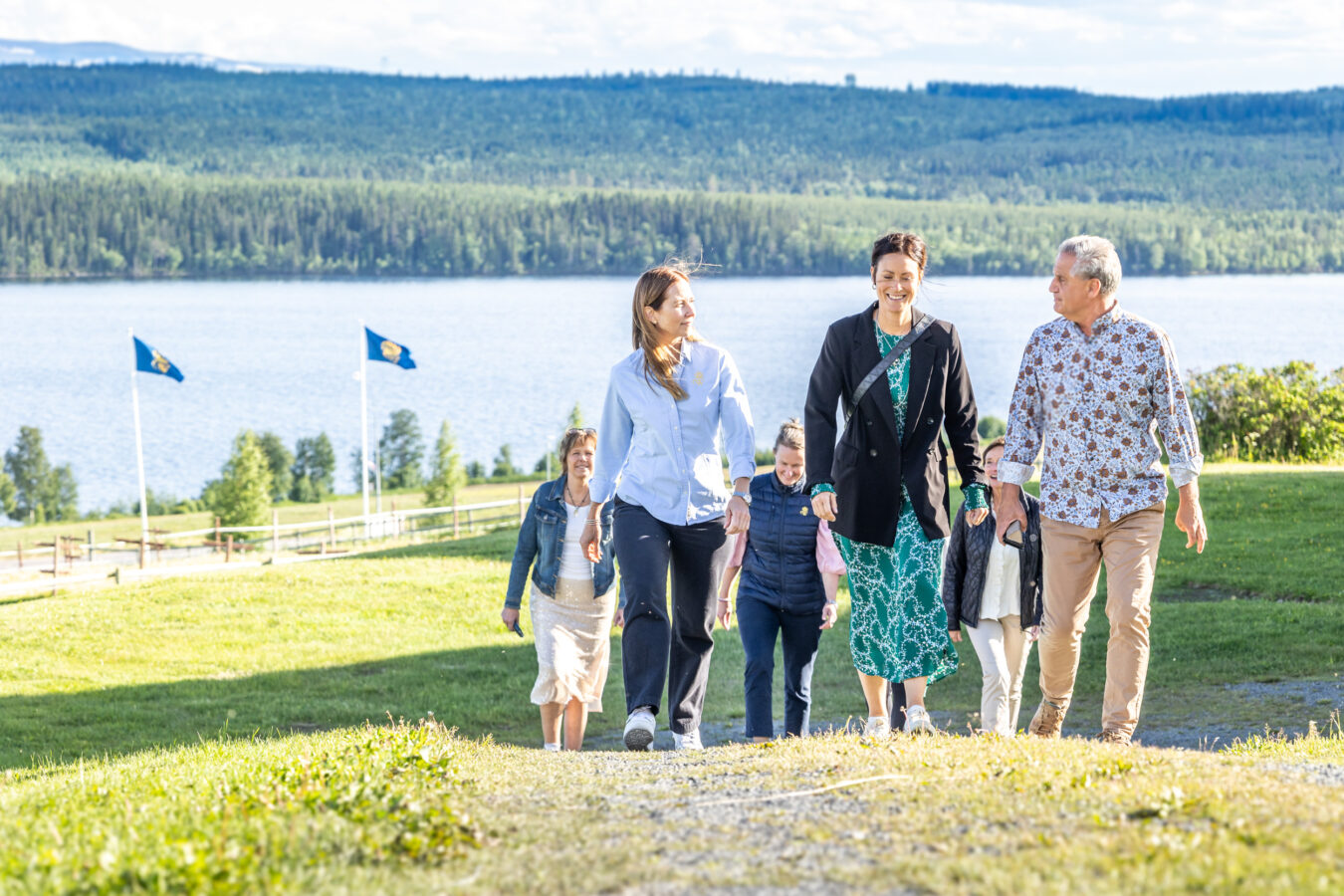 En grupp människor går uppför en gräsbevuxen kulle med en naturskön utsikt över sjön och skogen i bakgrunden. Flaggor med Wångens sigill syns till vänster.