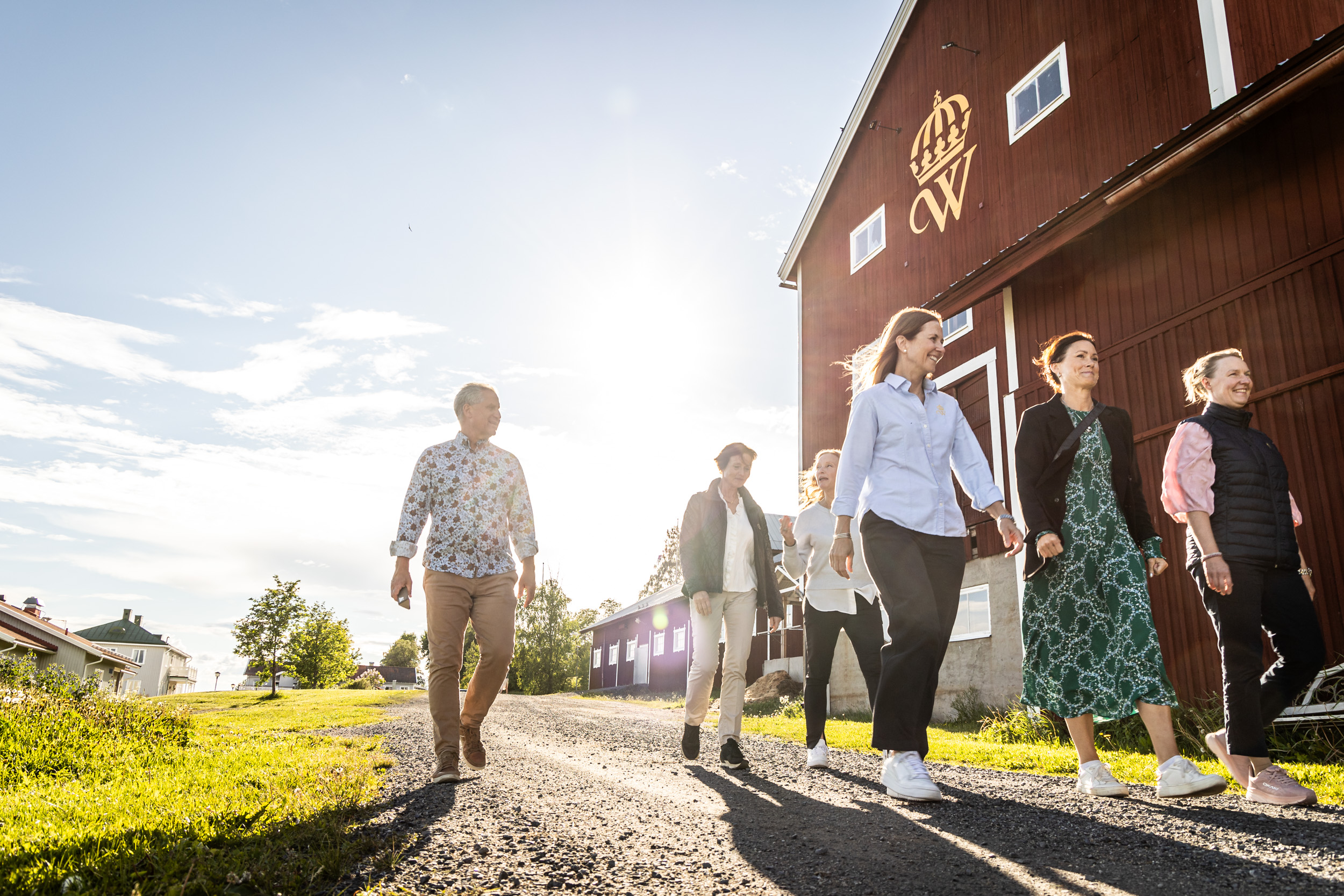 En grupp människor går på en grusgång bredvid en röd lada under en solig himmel.