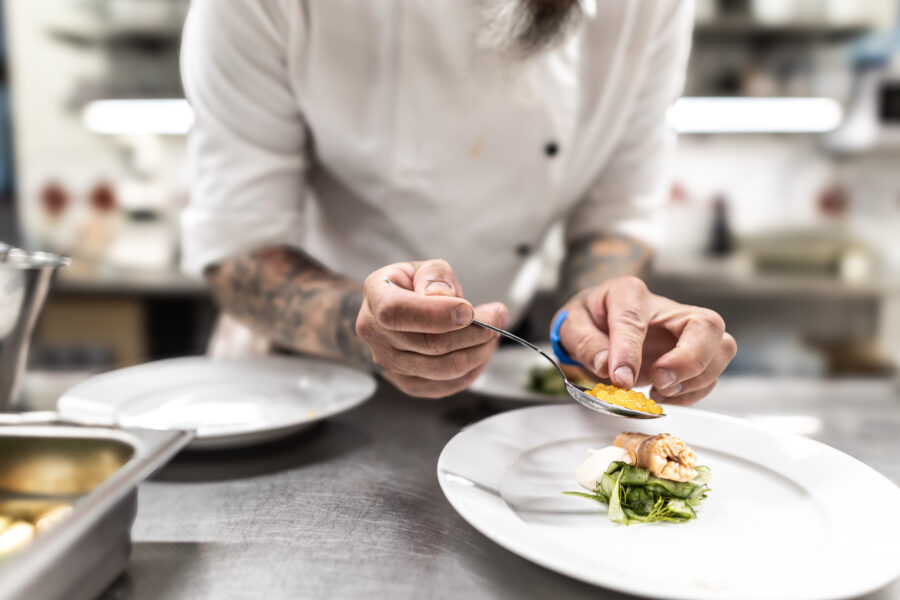 I ett livligt restaurangkök serverar en kock i en orörd vit uniform skickligt mat med en sked.