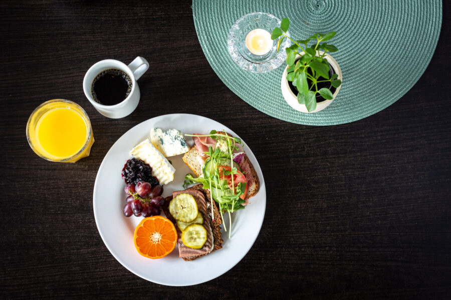 En frukosttallrik med rostat bröd, ostar, bär, vindruvor och skivad frukt bredvid glas apelsinjuice och kaffe, placerad på ett mörkt bord med en grön bordstablett och en krukväxt.