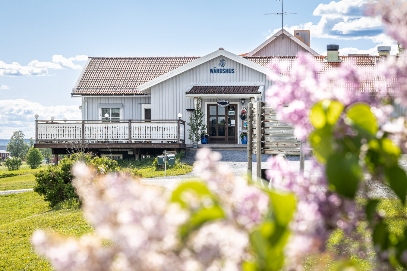 En vit träbyggnad med skylt, platsen är vackert accentuerad av grönska och rosa blommor i förgrunden, allt under en halvmolnig himmel.